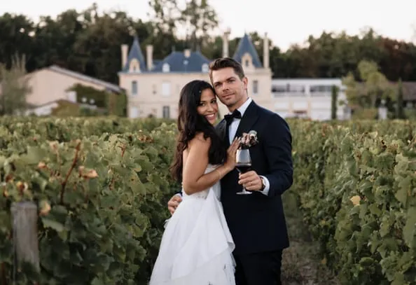 Couple de mariés tenant un verre de vin entre les rangs de vigne devant une demeure.