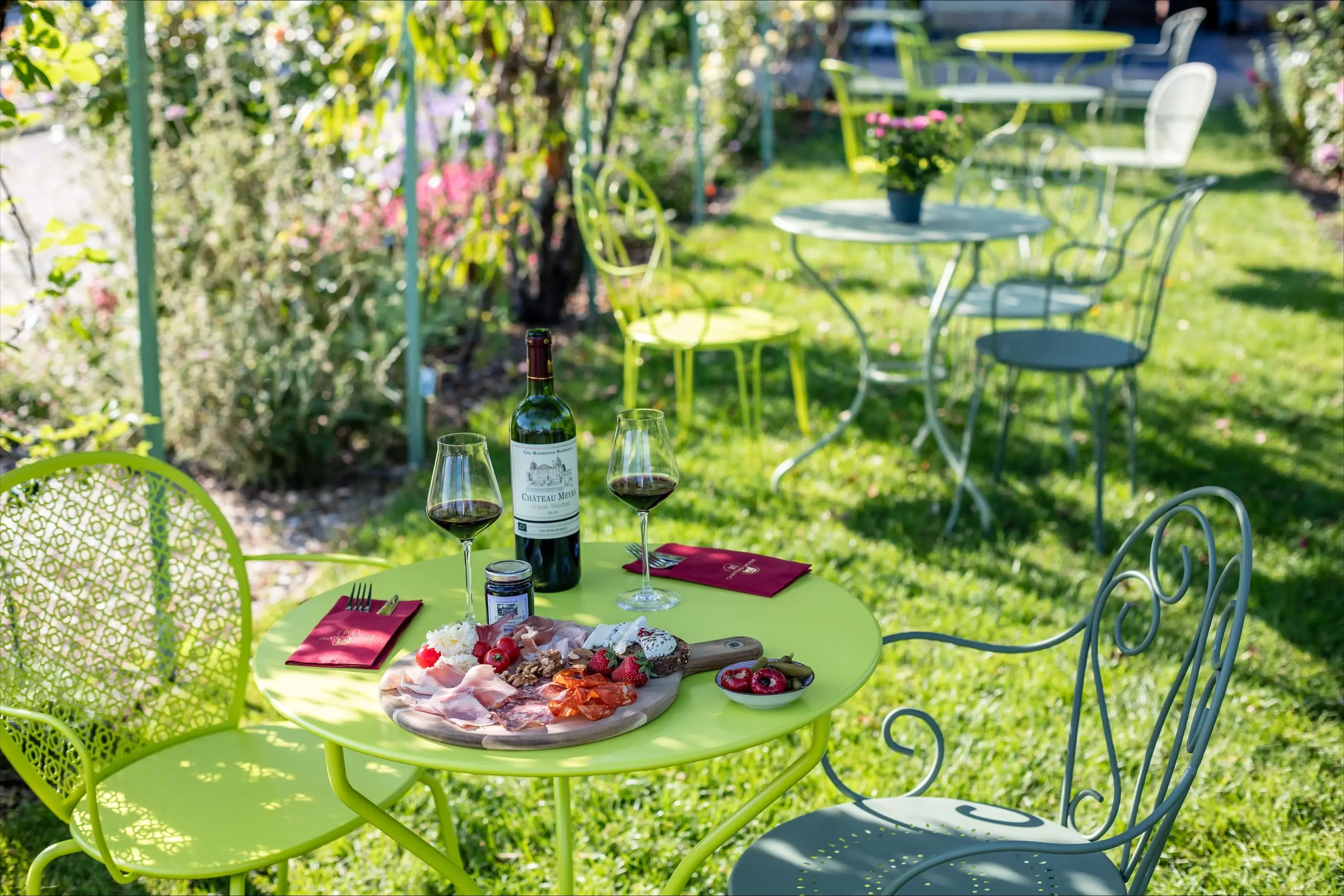 Table de jardin vert anis dressée d’une planche de charcuteries et de fromages, de deux verres de rouge et d’une bouteille de Château Meyre Haut-Médoc.